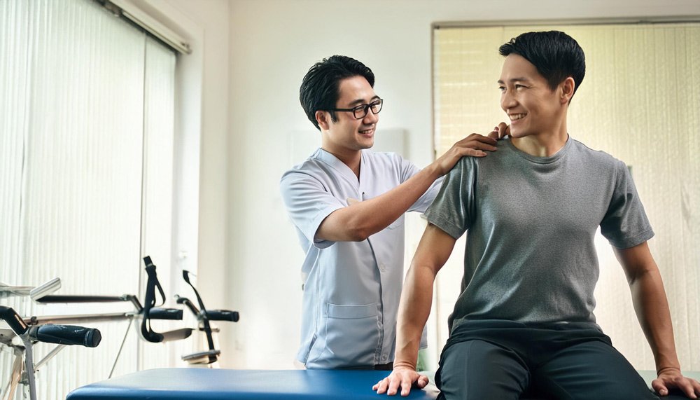 Physical therapist assisting a smiling male patient with shoulder exercises in a clinic setting, featuring therapy equipment in the background, designed for healthcare website design and physical therapy website, highlighting personalized rehabilitation and care.